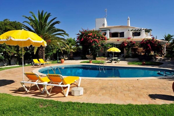 A pool area with lounge chairs and umbrellas, a swimming pool, and a building with a white facade and red flowers in the background.