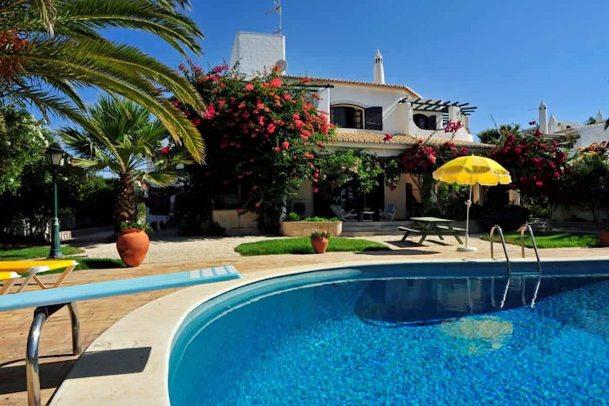 A swimming pool with a clear blue water, surrounded by a garden with palm trees and flowering plants.