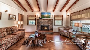 A spacious living room with a stone fireplace, wooden beams, and patterned furniture. - Bluebird View | Sierra Blanca Views + Hot Tub Retreat (Ruidoso)