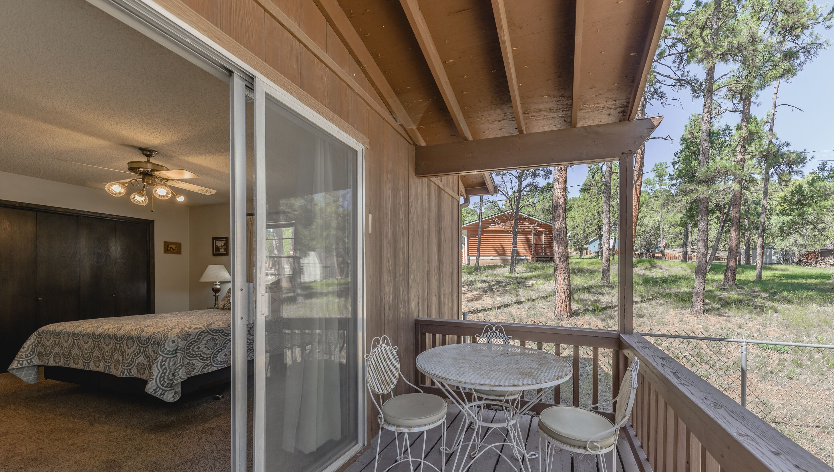 Un dormitorio con cama y ventilador de techo, un balcón con mesa y sillas, y vistas a una cabaña de madera y árboles.