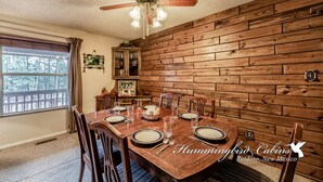Un comedor con una mesa de madera dispuesta para cuatro personas, un ventilador de techo y una ventana con vistas a árboles.