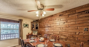 Un coin repas avec une table en bois dressée pour quatre personnes, un ventilateur de plafond et une fenêtre donnant sur les arbres.