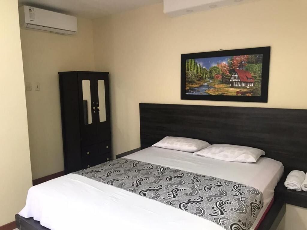 Una habitación de hotel con una cama, un cabecero oscuro, un cuadro enmarco de un paisaje, un armario y un aire acondicionado instalado en la pared.