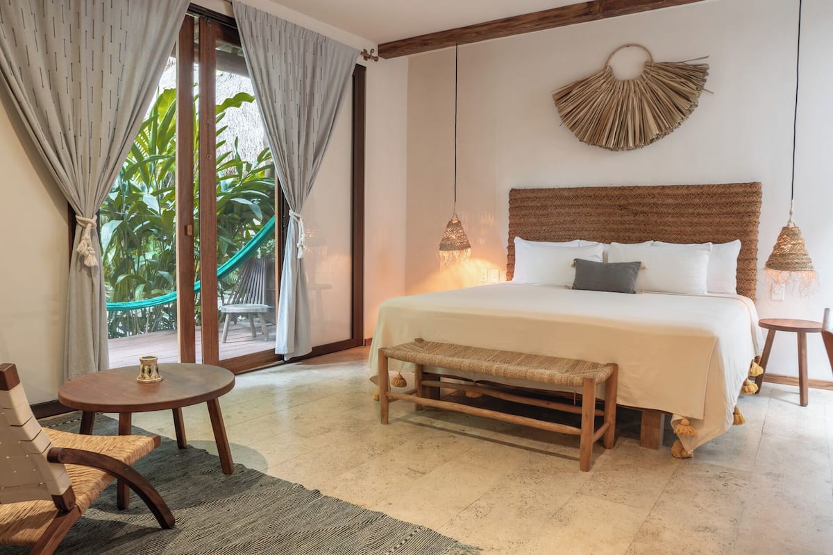 Une chambre à coucher comprenant un lit, deux tables de chevet, un banc, une chaise et une vue sur la verdure à l’extérieur.