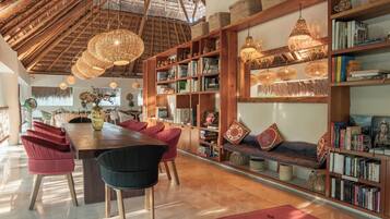 A spacious room with a high ceiling, wooden bookshelves, a dining area with a long table and chairs, and hanging wicker lamps.