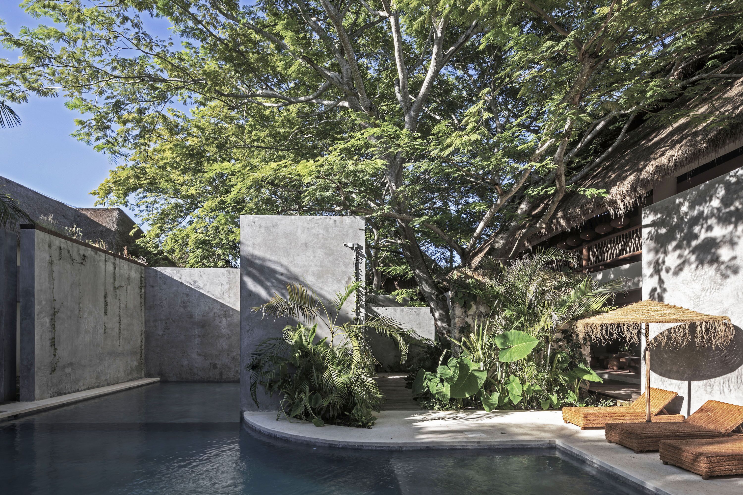 Un área de piscina con un muro de concreto, una sombrilla de paja y sillas de descanso.