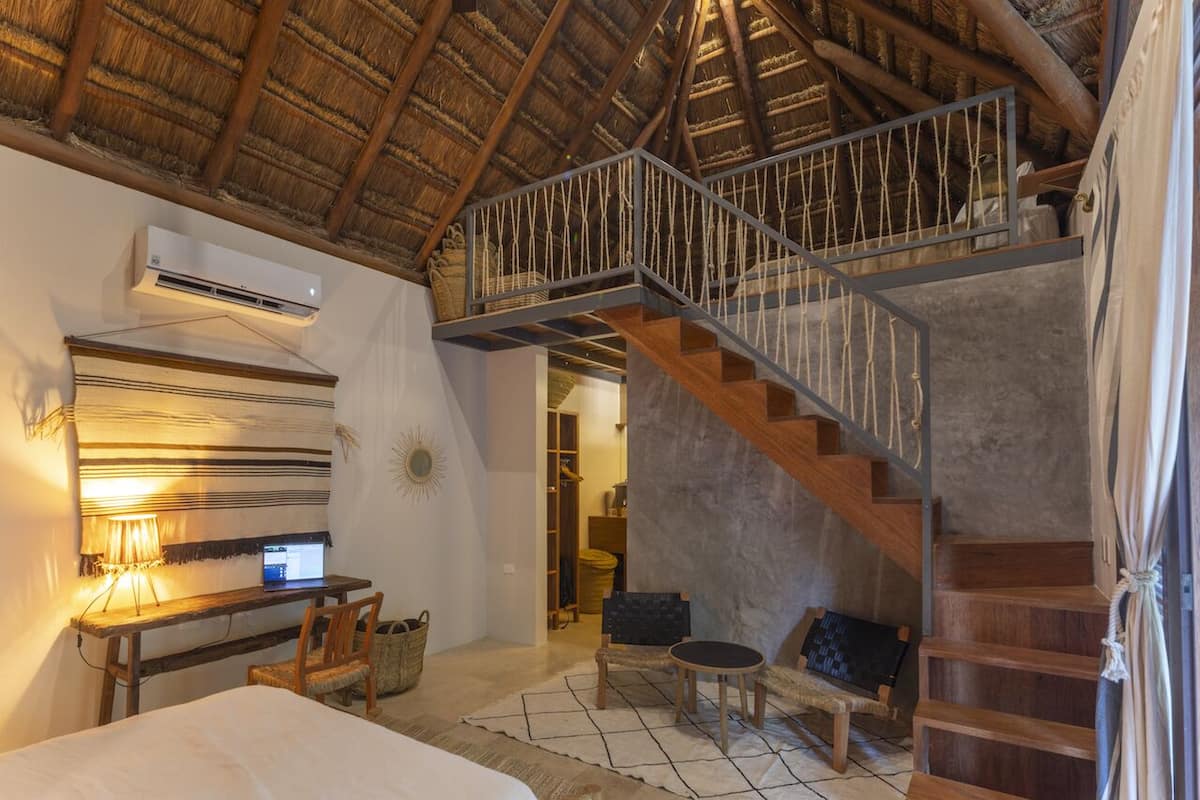 Une pièce rustique avec un toit de chaume, un escalier en bois, un bureau avec un ordinateur portable, un lit et un petit coin salon.