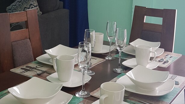 Un tavolo da pranzo apparecchiato con piatti, ciotole e bicchieri bianchi, con una parete verde e tende blu sullo sfondo.