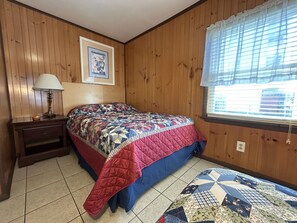 Ein Schlafzimmer mit Holzvertäfelung, einem Bett mit Quilzdecke, einem Nachttisch mit Lampe, einem Fenster mit Jalousien und einem gerahmten Bild an der Wand.
