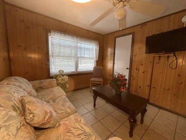 A room with wooden walls, a ceiling fan, a bed, a table with a vase of flowers, and a television mounted on the wall.