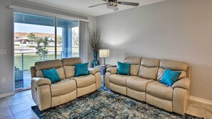 A living room with two beige sofas, a ceiling fan, a large window, and a view of a residential area. - Kissimmee Condo w/ Resort-Style Amenities! (Kissimmee)