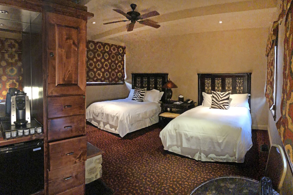 Habitación de hotel con dos camas, ventilador de techo, minibar y un espejo grande.