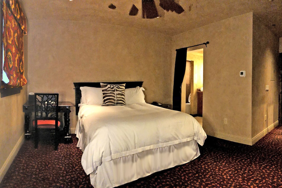 Habitación de hotel con una cama grande, una mesita de noche, una silla y una puerta que da a otra habitación.