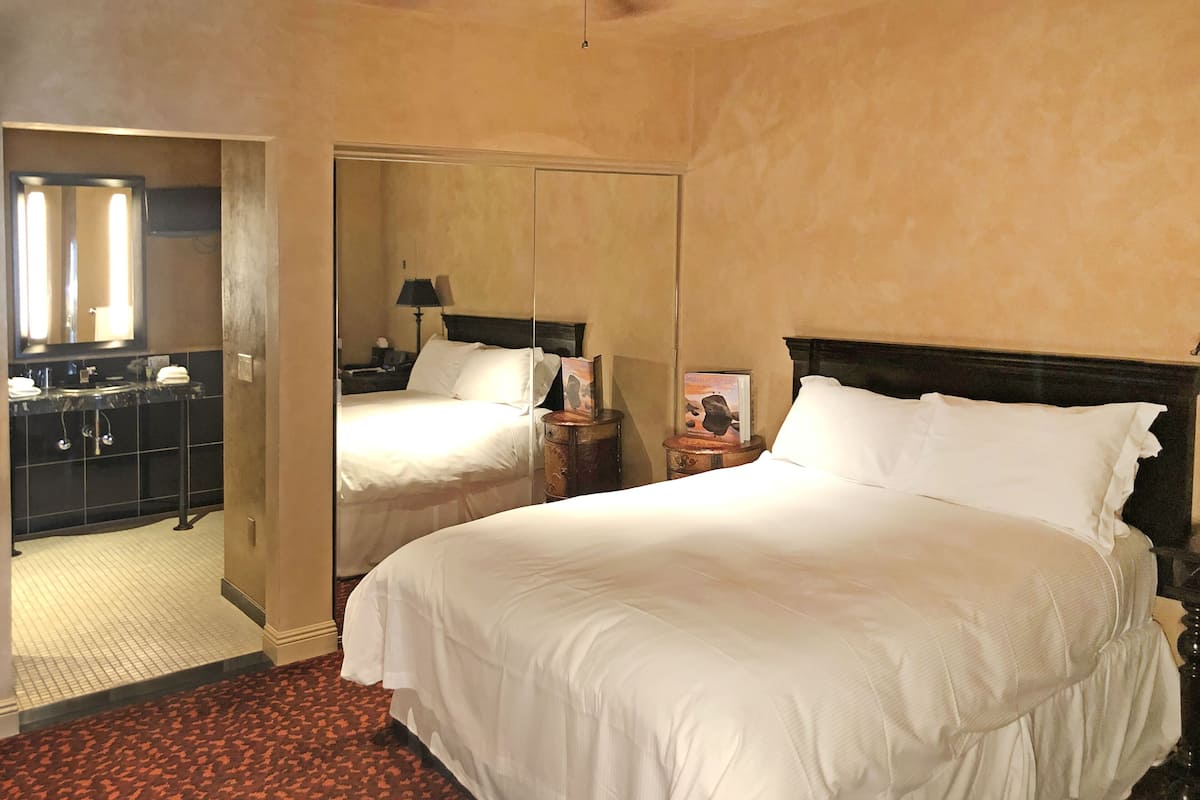 Habitación de hotel con una cama grande, un espejo, una mesita de noche y un baño con lavamanos y espejo.