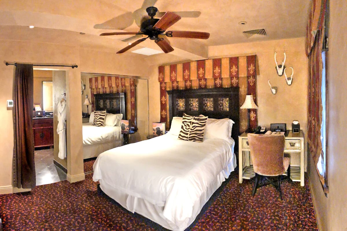 Una habitación de hotel con una cama grande, un ventilador de techo, un escritorio con una silla, un espejo y piso alfombrado.