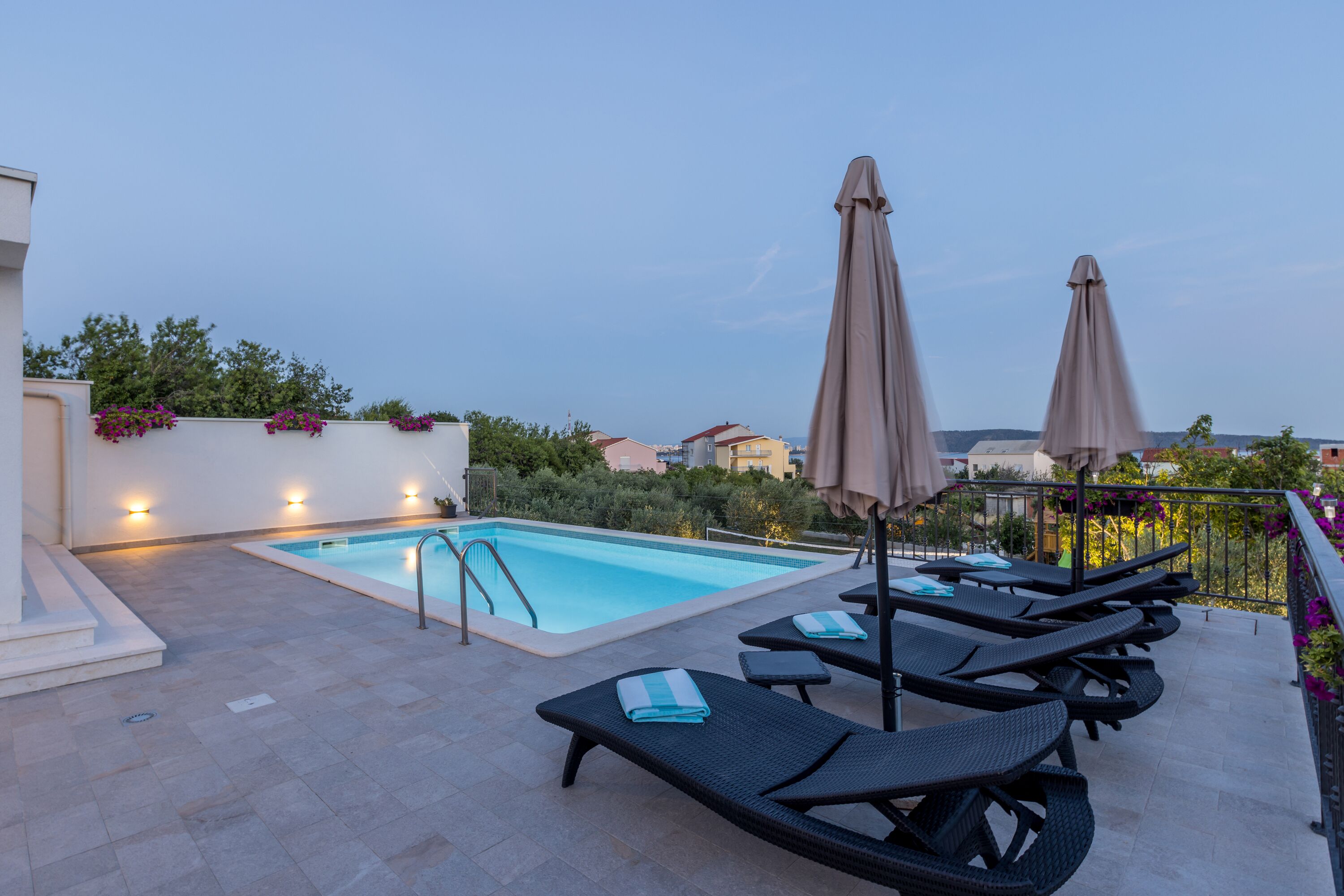 A rooftop pool area with lounge chairs and umbrellas, surrounded by greenery and buildings in the background.