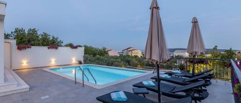 A rooftop pool area with lounge chairs and umbrellas, surrounded by greenery and buildings in the background.