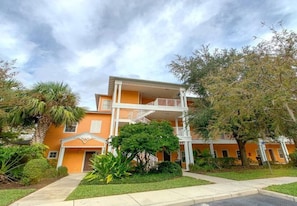 A multi-story building with a central balcony, surrounded by greenery and a clear sky.