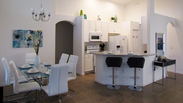 Une cuisine moderne avec des meubles blancs, un îlot central doté de tabourets de bar et un coin repas avec une table en verre et des chaises blanches.