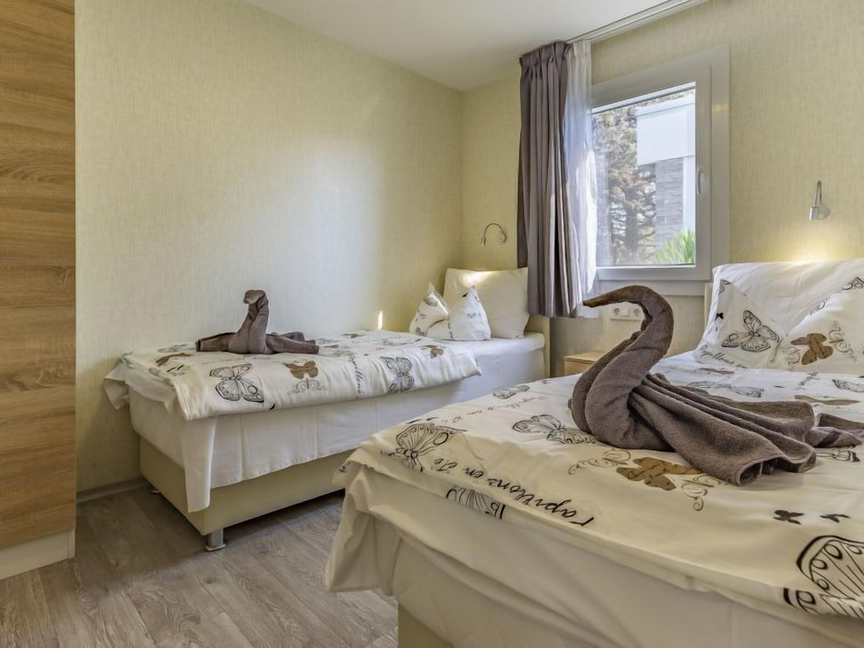 Une chambre avec deux lits, chacun agrémenté d’une serviette en forme de cygne. La chambre est dotée d’un plancher en bois, d’une fenêtre avec des rideaux et d’une table de chevet avec une lampe.