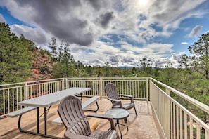 An outdoor deck with a picnic table and two Adirondack chairs, surrounded by a railing and overlooking a forested area.