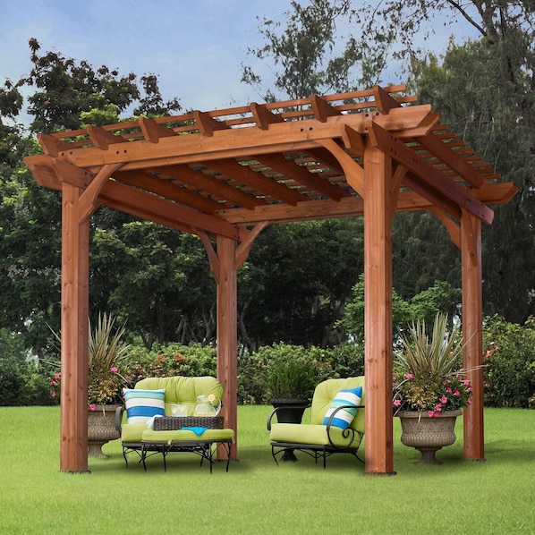 A wooden pergola structure with two green cushioned chairs and a small table, surrounded by lush greenery and potted plants.