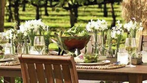 Une table à manger installée à l’extérieur, décorée de fleurs blanches dans des vases, avec un grand saladier au centre et des verres à vin.