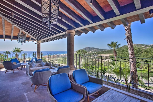 A covered outdoor seating area with wicker furniture and hanging lanterns, overlooking a coastal landscape.