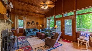 A cozy wooden interior with a fireplace, a blue sofa, and a large window overlooking greenery. - Rivers Edge in Todd - Home with hot tub- Enjoy the River from your front porch rocker (Todd)