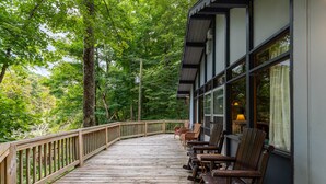 A wooden deck with chairs and a table, surrounded by trees. - Rivers Edge in Todd - Home with hot tub- Enjoy the River from your front porch rocker (Todd)