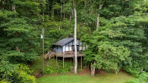 A cabin surrounded by dense forest, with a wooden deck and a small fire pit in the yard. - Rivers Edge in Todd - Home with hot tub- Enjoy the River from your front porch rocker (Todd)