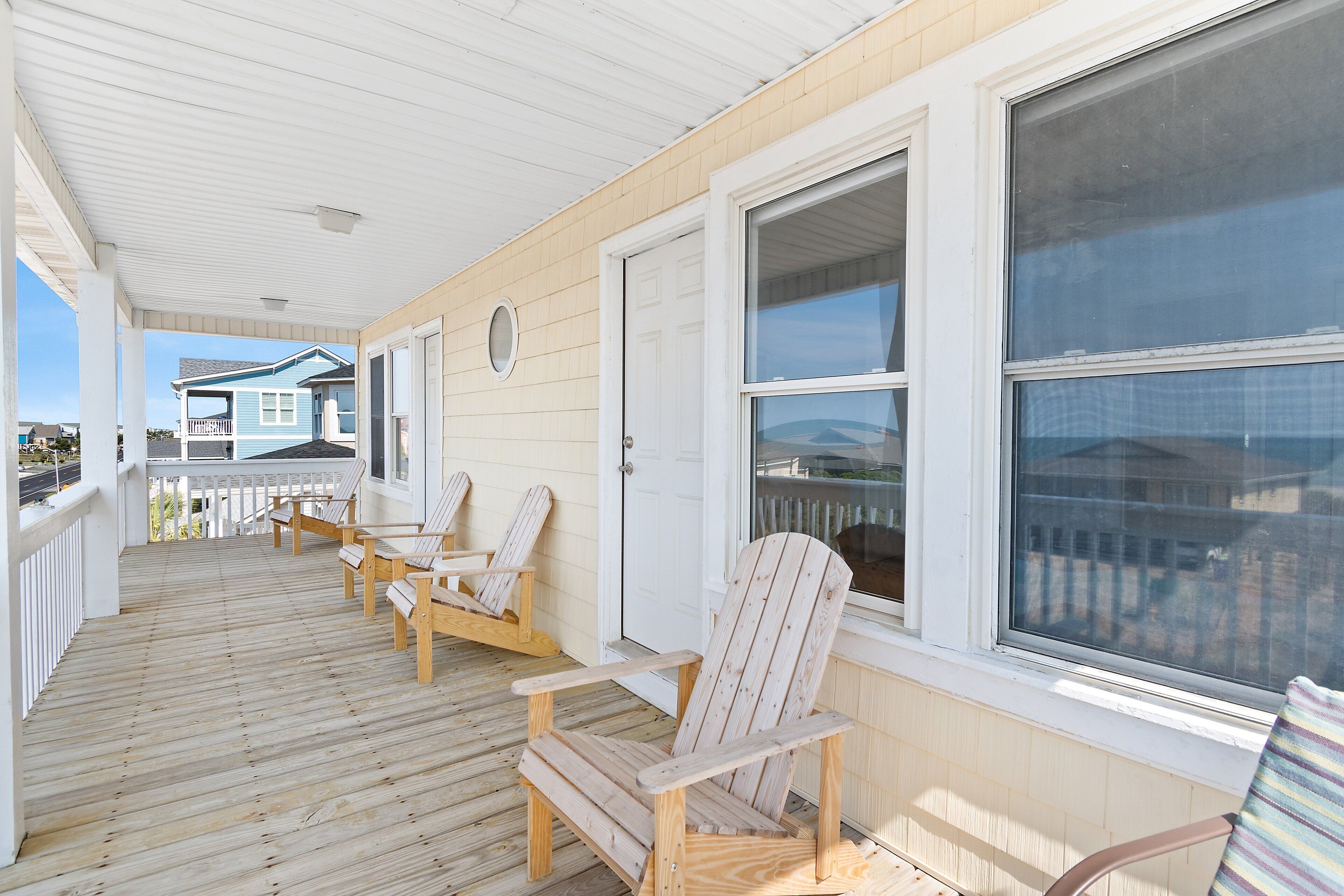 A wooden deck with four Adirondack chairs, a white door, and a large window.