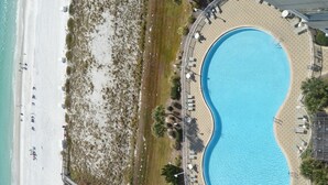 An aerial view of a resort with a large pool, beach, and a boardwalk with umbrellas and lounge chairs.