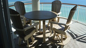 A round table with four Adirondack chairs on a balcony with a view of the ocean.