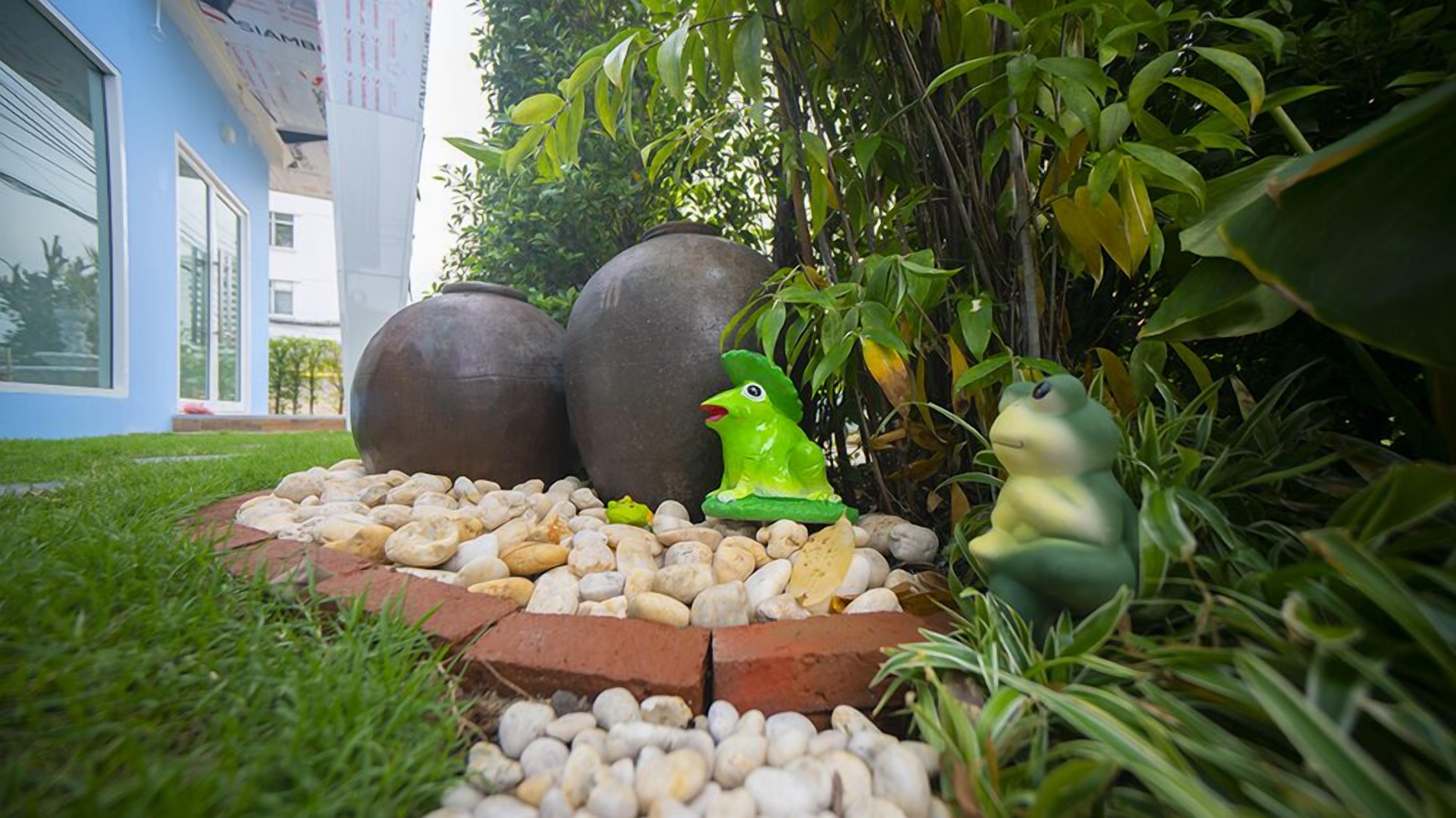 Un jardín con una estatua de rana, dos macetas grandes y un edificio al fondo.