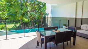 Un comedor con mesa y sillas de vidrio, ventilador de techo y vista a una piscina y follaje.