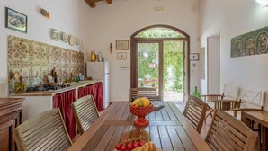 A dining area with a wooden table, chairs, and a fruit bowl. There is a kitchen area with a tiled backsplash and a refrigerator. - Beautiful private villa for 14 people with WIFI, private pool, TV, terrace and pets allowed (Buseto Palizzolo)