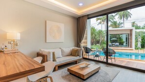 A modern living room with a wooden dining table, a sofa, and a coffee table. There is a large window with a view of a swimming pool and palm trees. - Zee Luxury Boutique Hotel (Ko Pha-ngan)