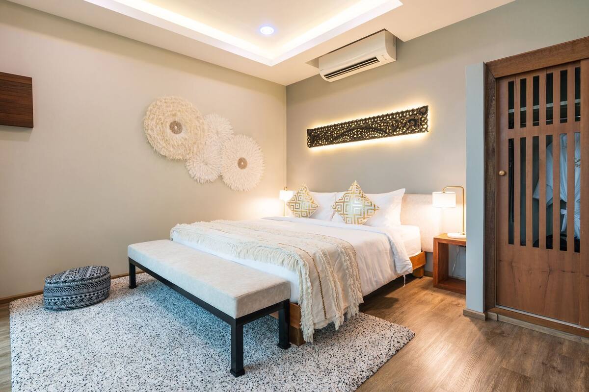 Un dormitorio moderno con una cama grande, mesitas de noche, un banco, un aire acondicionado de pared y una puerta de madera.