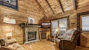Interior acogedor de una cabaña de troncos con chimenea de piedra, un sofá cómodo y un televisor montado encima.