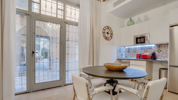 Een eetruimte met een ronde tafel, witte stoelen, een schaal, een klok en een keuken met apparatuur.
