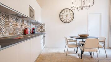 Une cuisine avec un mur recouvert de carreaux, une table ronde avec des chaises, une horloge et un lustre.