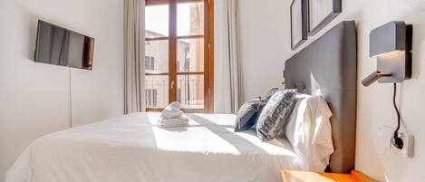 A hotel room with a large bed, a TV mounted on the wall, a window with curtains, and a bedside table with a lamp.