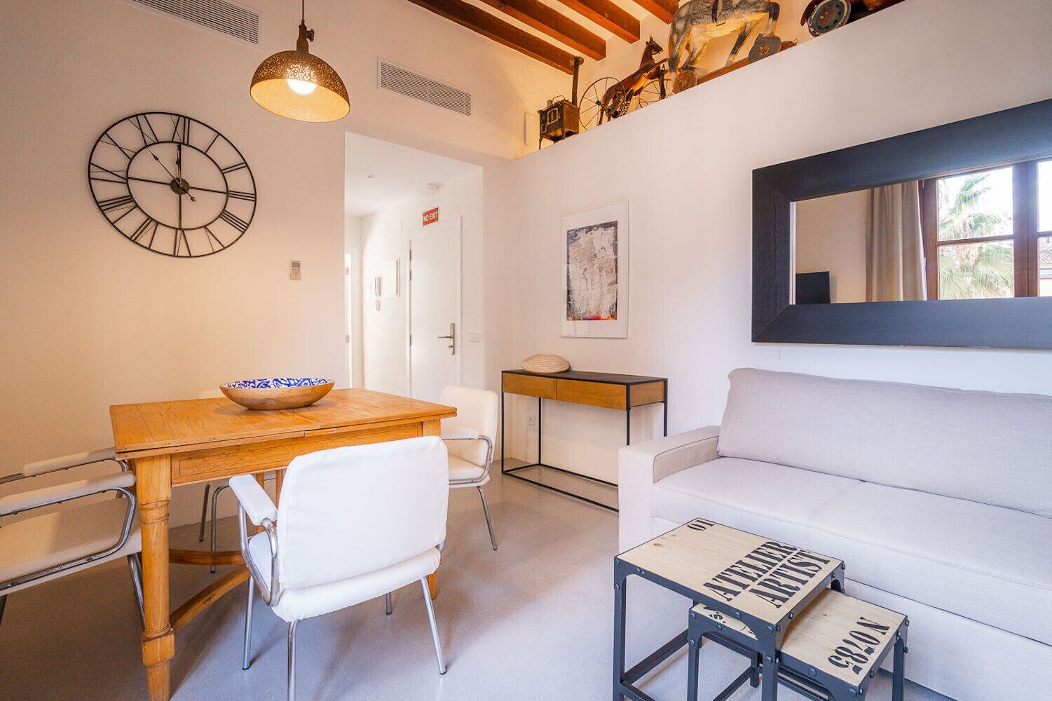 Un salon moderne avec une table à manger en bois, des chaises blanches, un canapé blanc, une petite table basse en bois, une horloge murale et un tableau abstrait encadré.