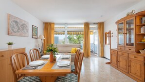 Un coin repas avec une table en bois dressée pour quatre personnes, des chaises avec des coussins à motifs, un canapé avec des coussins jaunes et un buffet en bois avec des portes en verre.