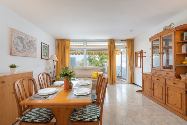 A dining area with a wooden table set for four, chairs with patterned cushions, a sofa with yellow cushions, and a wooden cabinet with glass doors.