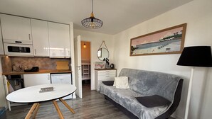 Une cuisine moderne avec des meubles blancs, une table à manger avec un plateau blanc et des pieds en bois, un canapé gris avec un coussin et une lampe sur pied.