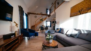 A spacious living area with a wooden floor, a grey sofa, a blue armchair, a wooden coffee table with a flower arrangement, and a staircase leading to an upper level.