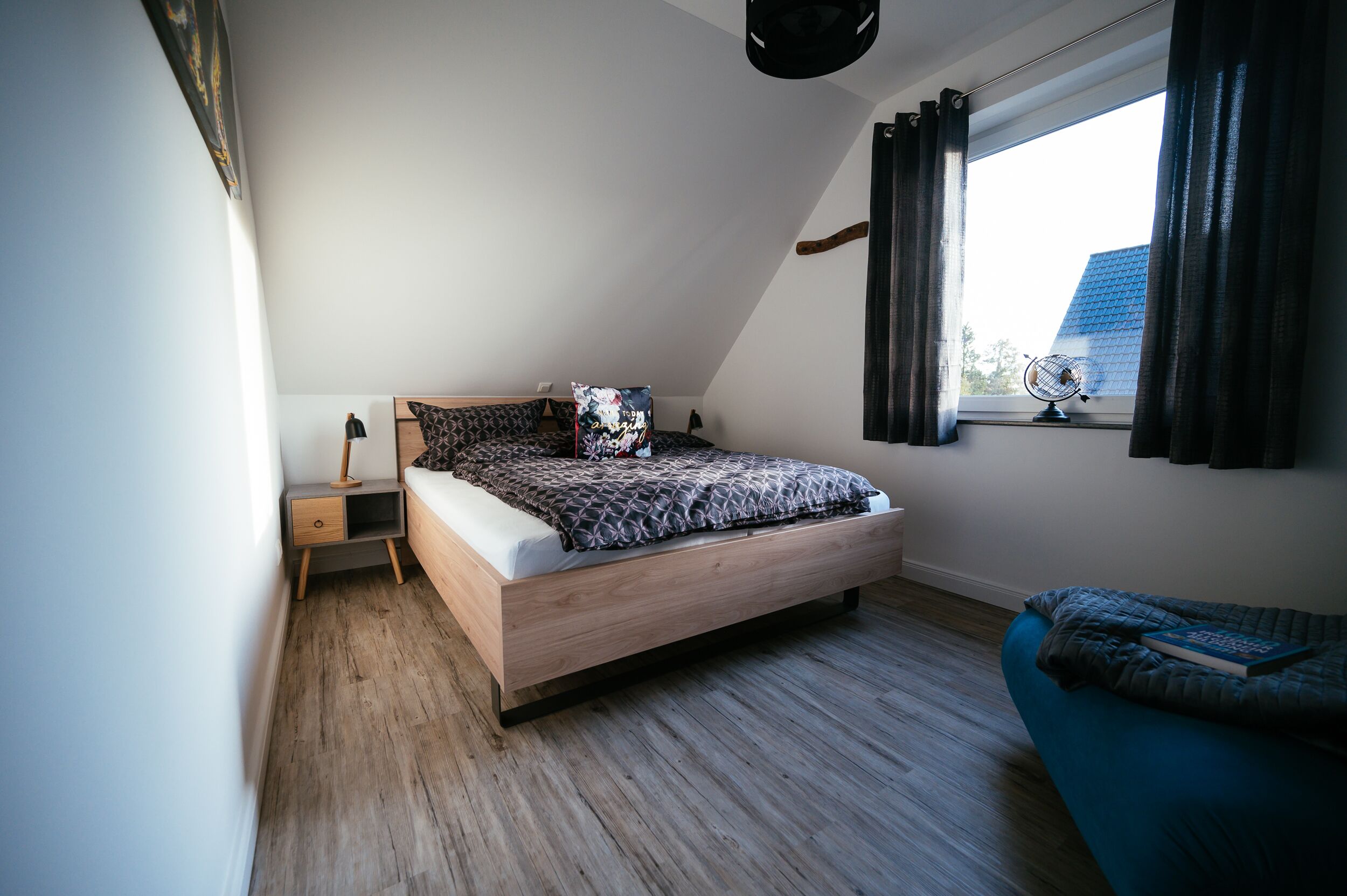 Een moderne slaapkamer met een schuine kap, een bed met beddengoed met een patroon, een nachtkastje met een lamp en een raam met gordijnen.