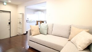 A modern living room with a beige sofa, a small rug, and a door leading to a kitchen area. - Xiaoningge (Osaka)